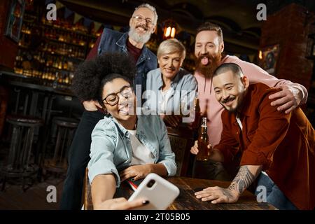 funny african american woman sticking out tongue and taking selfie with multiethnic workmates in bar Stock Photo