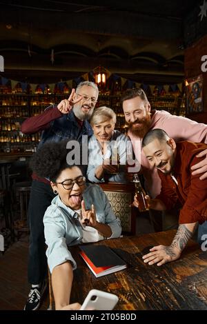 african american woman showing rock sign while taking selfie with multiethnic workmates in pub Stock Photo