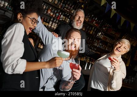overjoyed multiethnic colleagues with cocktails laughing in bar, diverse team having fun after work Stock Photo