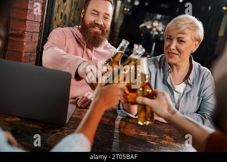 cheerful bearded man and middle aged woman clinking beer bottles with colleagues near laptop in pub Stock Photo