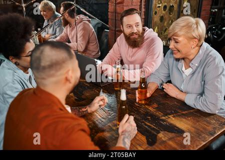 middle aged woman laughing near multiethnic colleagues next to laptop and beer bottles in pub Stock Photo