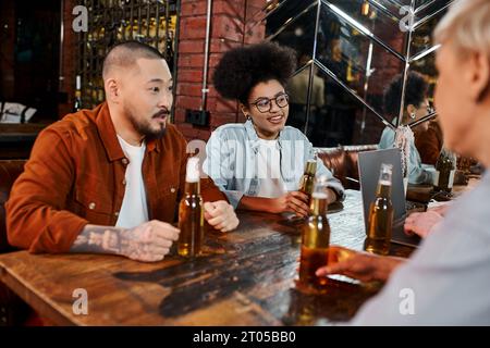 happy diverse team of multiethnic workmates discussing startup near beer bottles and laptop in pub Stock Photo
