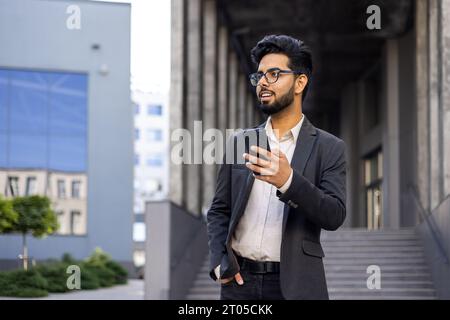 A young businessman in a business suit and glasses is walking around the city outside an office building, the man is holding a phone in his hands, typing a message and using an application. Stock Photo