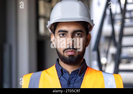 Close-up portrait of a young male engineer wearing a hard hat and vest, worker worker looking focused at the camera, working in an industrial plant. Stock Photo
