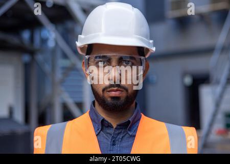 Close-up portrait of a young male engineer wearing a hard hat and vest, worker worker looking focused at the camera, working in an industrial plant. Stock Photo