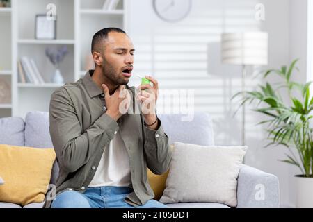 African american young man sitting on sofa at home and blowing throat with medicine spray, suffering from pain and illness. Stock Photo