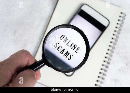 A man's hand holds a black magnifying glass by the handle on a smartphone background with online scams text. Stock Photo