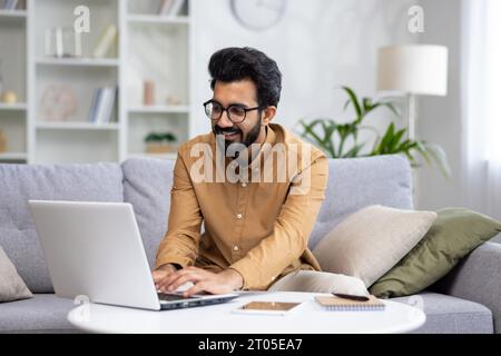 Young Indian male freelancer working online at home using laptop. He sits smilingly on the sofa and types on the keyboard, corresponds, executes the project. Stock Photo