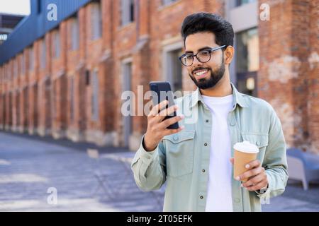 A young student uses the phone, the man holds a smartphone and a cup of hot drink in his hands, walks around the city, smiles, reads messages and watches online videos. Stock Photo