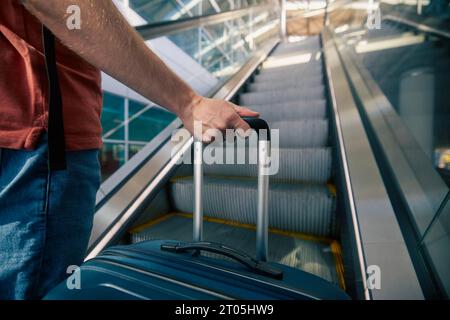 Traveling by airplane. Man standing on escalator in airport terminal. Selective focus on hand holding suitcase. Passenger is ready for travel. Stock Photo