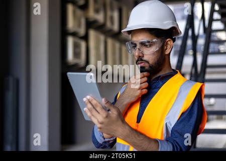 Serious thinking and focused engineer working in factory in hard hat and vest, man using tablet computer reading diagram, and checking equipment, inside industrial factory. Stock Photo
