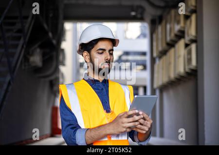 Serious thinking and focused engineer working in factory in hard hat and vest, man using tablet computer reading diagram, and checking equipment, inside industrial factory. Stock Photo