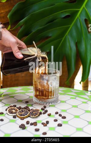 Hand pouring cold brew coffee in a bottle with transparent glass full of ice on green leaves background Stock Photo