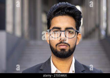 Close up portrait of young successful businessman, thinking serious concentrated man looking at camera outside office building, wearing suit and glasses. Stock Photo