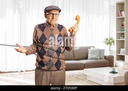 Confused elderly man standing tangled in a cable from a vintage rotary phone in a living room Stock Photo