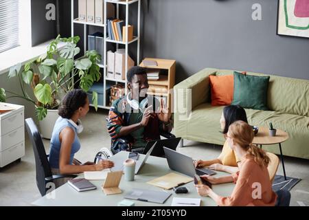 High angle portrait of animated young Black man with disability sharing ideas with colleagues during business meeting and smiling Stock Photo
