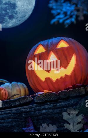 A smiling Jack O Lantern sitting on an old wooden porch with small pumpkins and fallen leaves as a Halloween decoration. With a full moon above. Stock Photo