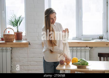 A woman in the kitchen puts vegetables from a rope bag on the table Stock Photo