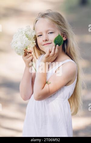 Portrait of a young beautiful girl holding hydrangea flowers and a green leaf in front of her face, smiling. Vertical frame. Stock Photo