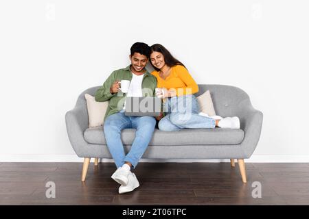 Loving young indian couple in casual outfit sitting on sofa over white wall background, using laptop and drinking coffee Stock Photo