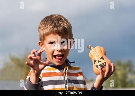 Portrait of cute boy holding a small jack lantern in his hands and grimacing. Background blue sky, bright october day. happy halloween. Stock Photo