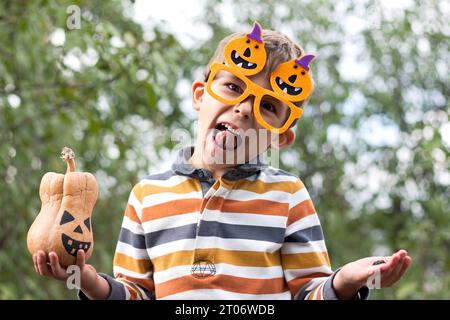 Portrait of cute boy in funny orange glasses holding a little jack lantern in his hands and grimacing in park. Happy Halloween. Stock Photo