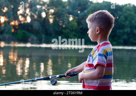 Portrait of boy with fishing rod in profile. Focused young fisherman is fishing in river on summer day. Amateur fishing in open water from river bank. Stock Photo