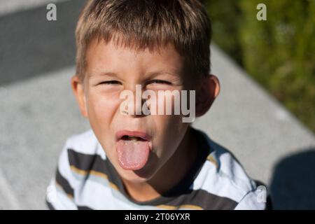Close-up of child shows tongue. Irritated boy is teased and stuck out his tongue. Portrait of boy with grimace on his face in park on summer sunny day Stock Photo