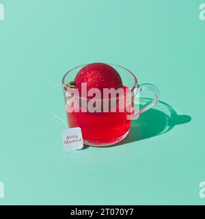 Creative Christmas composition of transparent glass cup of fruit tea with teabag made of red Christmas bauble on green background. Minimal concept. Stock Photo