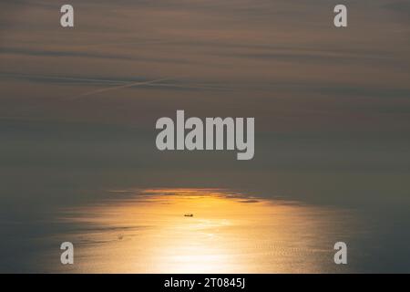 A ship in the middle of the ocean lit up by orange sunlight on water Stock Photo