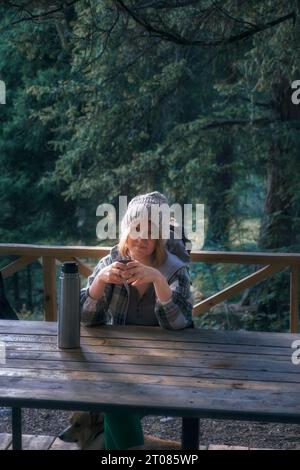 A woman tourist backpacker drinks tea from a thermos in nature in a mountain autumn forest at a wooden table Stock Photo