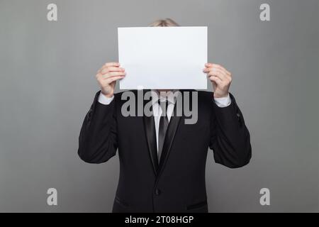 Anonymous businessman hiding his head behind blank empty white paper against gray studio wall background Stock Photo