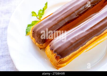 French pastry eclairs with cream filling covered with melted milk chocolate and mint leaves. Eclairs with custard and chocolate icing on a white plate Stock Photo
