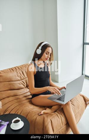 barefoot freelancer in wireless headphones using laptop and sitting on bean bag chair, pretty woman Stock Photo