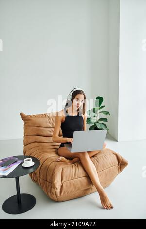 pretty freelancer in wireless headphones using laptop and sitting on bean bag chair, young woman Stock Photo