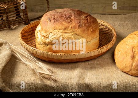 A loaf of homemade white bread in a basket on a burlap tablecloth. Nearby lies a bunch of barley ears Stock Photo