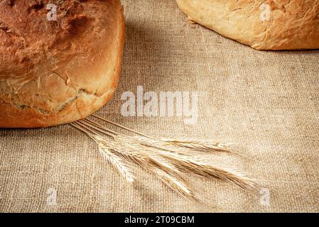 The corners of loaves of white bread and a bunch of barley ears are visible on the burlap tablecloth. Stock Photo
