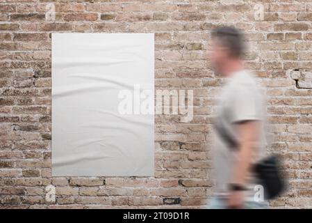 Wrinkled, blank A3 poster in white, adhered to a brick wall. A man walks beside. Ad or marketing campaign design promotion Stock Photo