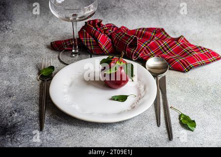 High angle of ripe red apple in plate placed between knife,spoon and fork with cloth and empty glass on gray surface Stock Photo