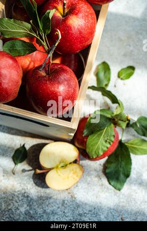 Top view of whole fresh red ripe organic delicious apples with green leaves filled in wooden box with half cut pieces and placed on gray surface in da Stock Photo