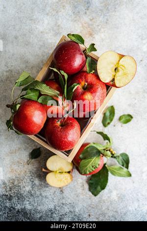 Top view of whole fresh red ripe organic delicious apples with green leaves filled in wooden box with half cut pieces and placed on gray surface in da Stock Photo