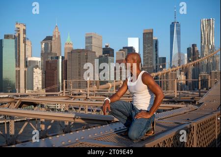 African American male sitting at metal suspension Brooklyn bridge infrastructure and looking away in daylight cityscape skyscrapers of Manhattan with Stock Photo