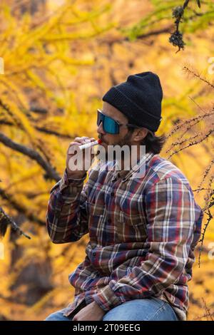 Positive man sitting on forest in autumn with a cap on his head and a cigarette in his mouth while resting against a blurred background Stock Photo