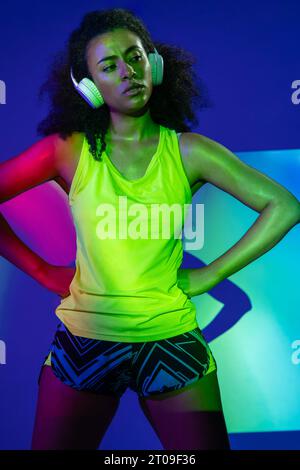 Young slim African American female in activewear standing with hands on waist and listening to songs on headphones against purple background looking a Stock Photo