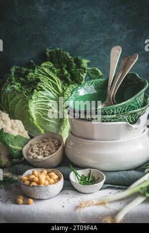 From above decorated table with various dishware ceramic bowls and spoons with fresh cauliflower, cabbage, legumes and herbs placed on table against g Stock Photo