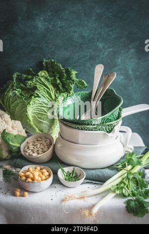 From above decorated table with various dishware ceramic bowls and spoons with fresh cauliflower, cabbage, legumes and herbs placed on table against g Stock Photo