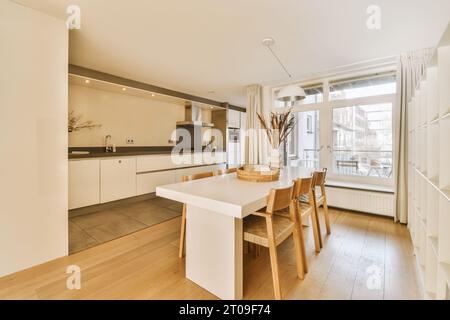 Interior of open plan kitchen with white dining table and wooden chairs next to window at contemporary home Stock Photo