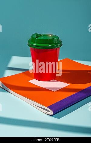 Minimalist red plastic cup of coffee and green lid placed on table on top of notebook against blue background Stock Photo