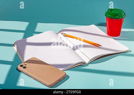 Minimalist red plastic cup of coffee and green lid along with a notebook and a mobile phone against blue background Stock Photo
