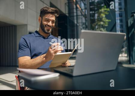 Smiling businessman working on laptop sitting in office background and making notes Stock Photo